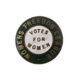 A vintage circular badge with a green outer ring and a white inner circle containing the words 'VOTES FOR WOMEN.' The outer ring features the text 'WOMENS FREEDOM LEAGUE.' The badge appears to be from a historical movement advocating for women's voting rights.