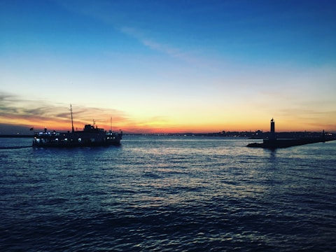 A serene cruise ship sailing past a glowing lighthouse at sunset.