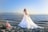 A bride in a flowing white wedding gown stands gracefully on a sandy beach, with the ocean and distant mountains in the background. A bouquet of pink and white flowers lies on the sand nearby under the clear blue sky.