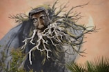 A mural featuring a surreal image of a face with deep textures and expressive eyes, integrated into what appears to be a combination of branches and roots. The organic elements give the impression of transformation or a blend of humanity and nature, with a muted earthy background enhancing the artwork.