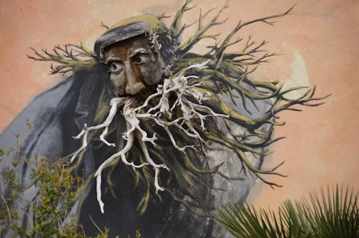 A mural featuring a surreal image of a face with deep textures and expressive eyes, integrated into what appears to be a combination of branches and roots. The organic elements give the impression of transformation or a blend of humanity and nature, with a muted earthy background enhancing the artwork.