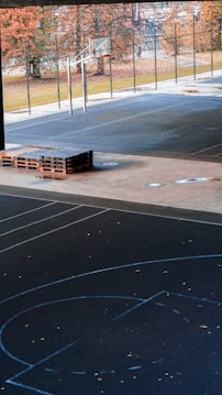 An outdoor basketball court with a hoop and backboard surrounded by a tall fence. The ground is a dark asphalt surface with blue court markings. There are wooden pallets stacked on the side of the court, and autumnal trees with orange leaves are visible in the background.