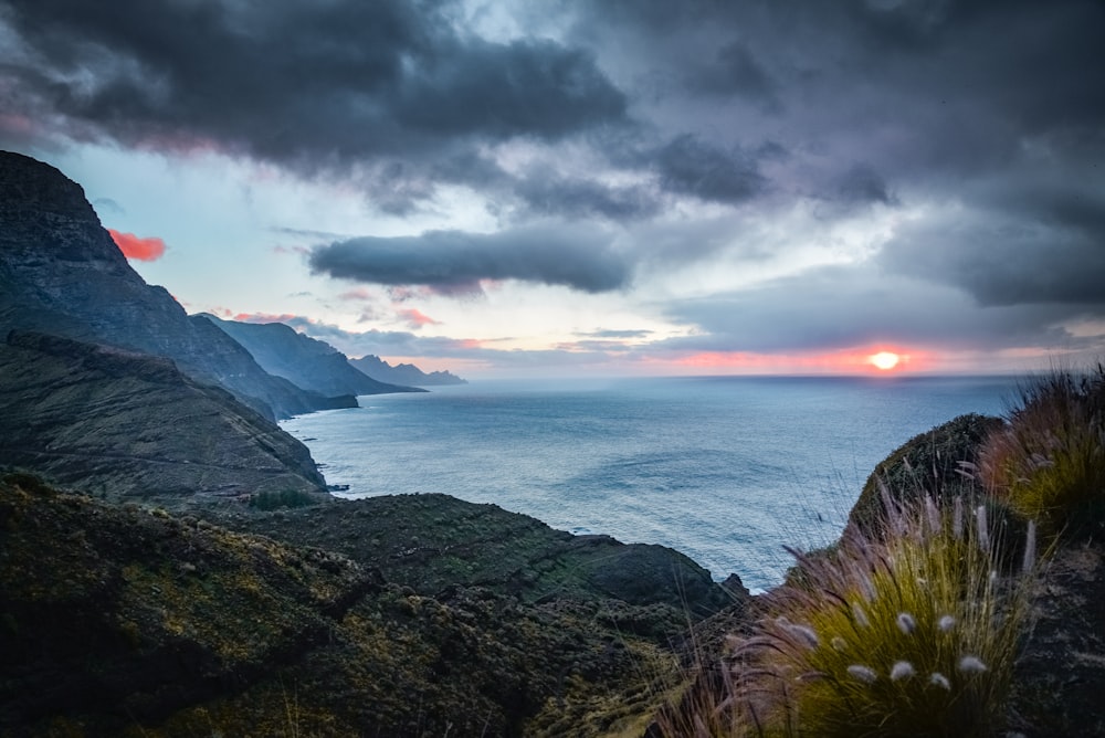 green and brown mountain beside sea under gray clouds during sunset