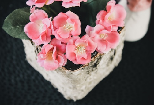 A collection of vibrant pink flowers, possibly artificial, arranged in a decorative pot with textured bark-like accents. The background is a textured dark surface, enhancing the contrast with the colorful floral arrangement. Green leaves accompany the flowers, providing a natural look.