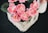 A collection of vibrant pink flowers, possibly artificial, arranged in a decorative pot with textured bark-like accents. The background is a textured dark surface, enhancing the contrast with the colorful floral arrangement. Green leaves accompany the flowers, providing a natural look.