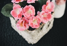 A collection of vibrant pink flowers, possibly artificial, arranged in a decorative pot with textured bark-like accents. The background is a textured dark surface, enhancing the contrast with the colorful floral arrangement. Green leaves accompany the flowers, providing a natural look.