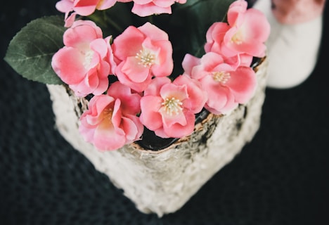 A collection of vibrant pink flowers, possibly artificial, arranged in a decorative pot with textured bark-like accents. The background is a textured dark surface, enhancing the contrast with the colorful floral arrangement. Green leaves accompany the flowers, providing a natural look.