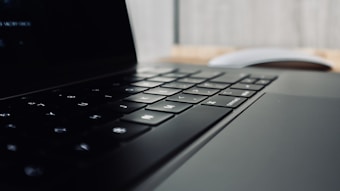 A close-up view of a laptop keyboard with blurred background, focusing on the keys and part of the trackpad. The lighting creates a sleek and modern appearance.