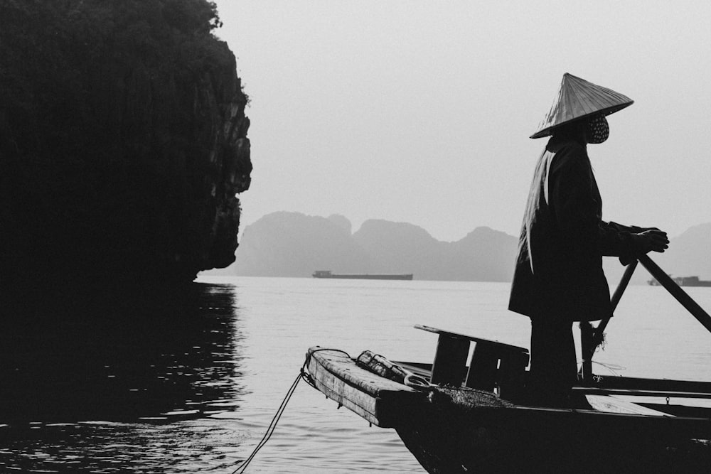 grayscale photo of man in black jacket and hat standing on boat on body of water