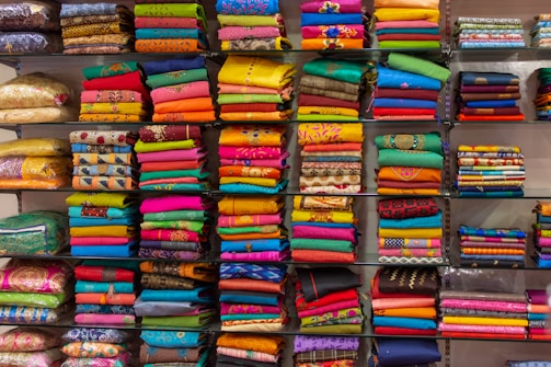 A display of colorful folded fabrics arranged neatly on shelves. The fabrics showcase a variety of patterns and textures, ranging from bright pinks, greens, and blues to intricate embroidery and designs.