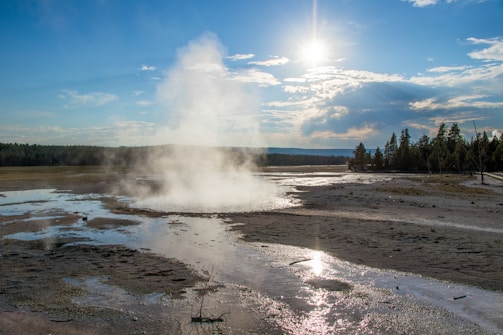 A landscape with a geothermal hot spring expelling steam into the air. The ground is wet with mineral deposits and the surrounding area is largely flat with trees along the horizon. The sky is partly cloudy with the sun shining brightly, reflecting off the water on the ground.