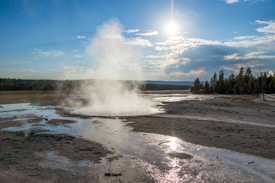 A landscape with a geothermal hot spring expelling steam into the air. The ground is wet with mineral deposits and the surrounding area is largely flat with trees along the horizon. The sky is partly cloudy with the sun shining brightly, reflecting off the water on the ground.