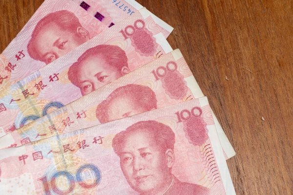 Several 100 Yuan Chinese banknotes are fanned out on a wooden surface. The notes are predominantly red and feature intricate designs and Chinese characters.