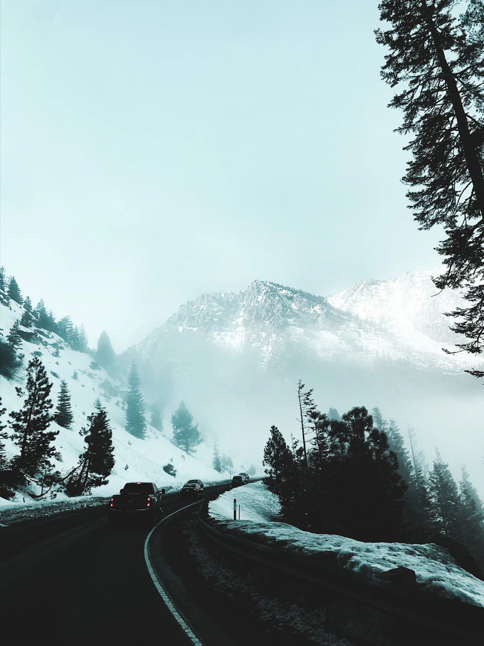 The image depicts a winding road cutting through a snowy mountain range. The sky above is overcast, casting a soft light on the scene below. Two cars are visible on the road, one driving towards the camera and another moving away from it. The mountains in the background are blanketed in snow with patches of green trees scattered throughout.
