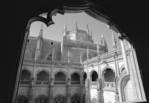 A black and white photograph captures an intricately designed Gothic-style courtyard. The architecture features pointed arches, ornate spires, and detailed stone carvings, with the image framed by a dark, arched window.