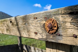 A close-up of a weathered wooden fence with visible rusting metal fixtures. The fence's texture is rough and aged, with natural grain patterns. A rusty circular dial is attached to the wood, showing signs of wear. The background features a clear blue sky and hints of greenery.