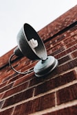 A close-up view of a metal exterior wall-mounted lamp attached to a brick wall, featuring a compact fluorescent light bulb. The lamp has a textured surface and curves elegantly, with the focus on its structure against the red bricks. The perspective is angled, giving a dynamic view of the lamp against the brick pattern.