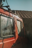 A red vehicle is being washed, with water droplets visible on the windows and body. Sunlight glints off the wet surface, creating a shimmering effect. The background shows a blurred rooftop.