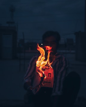 A person is sitting in a dimly lit environment, holding a burning newspaper. The flames create a striking contrast with the dark background, illuminating both the newspaper and the person's figure partially.