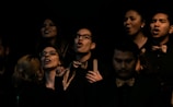 A group of people, mostly wearing dark attire with gold bow ties, are captured mid-song. Several individuals have their mouths open, likely singing, while others are smiling. The lighting is dim, creating a dramatic and intimate atmosphere. The focus is mainly on their expressions and attire.