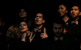 A group of people, mostly wearing dark attire with gold bow ties, are captured mid-song. Several individuals have their mouths open, likely singing, while others are smiling. The lighting is dim, creating a dramatic and intimate atmosphere. The focus is mainly on their expressions and attire.