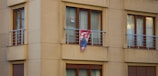 A beige apartment facade with multiple windows and a small balcony featuring metal railings. A colorful item, possibly a blanket or tapestry, with a superhero emblem is hanging over the balcony railing. The windows have brown wooden frames and white curtains. A small poster with a face is visible in one window.