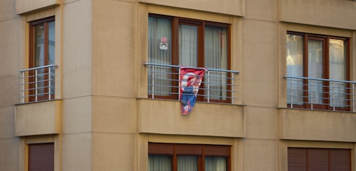 A beige apartment facade with multiple windows and a small balcony featuring metal railings. A colorful item, possibly a blanket or tapestry, with a superhero emblem is hanging over the balcony railing. The windows have brown wooden frames and white curtains. A small poster with a face is visible in one window.