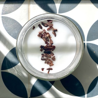 A top-down view of a jar filled with a white, creamy substance, likely yogurt or a dessert, topped with chocolate pieces. The jar is placed on a patterned surface featuring circular, black, and white geometric designs. Sunlight creates diagonal shadows across the composition, adding depth.