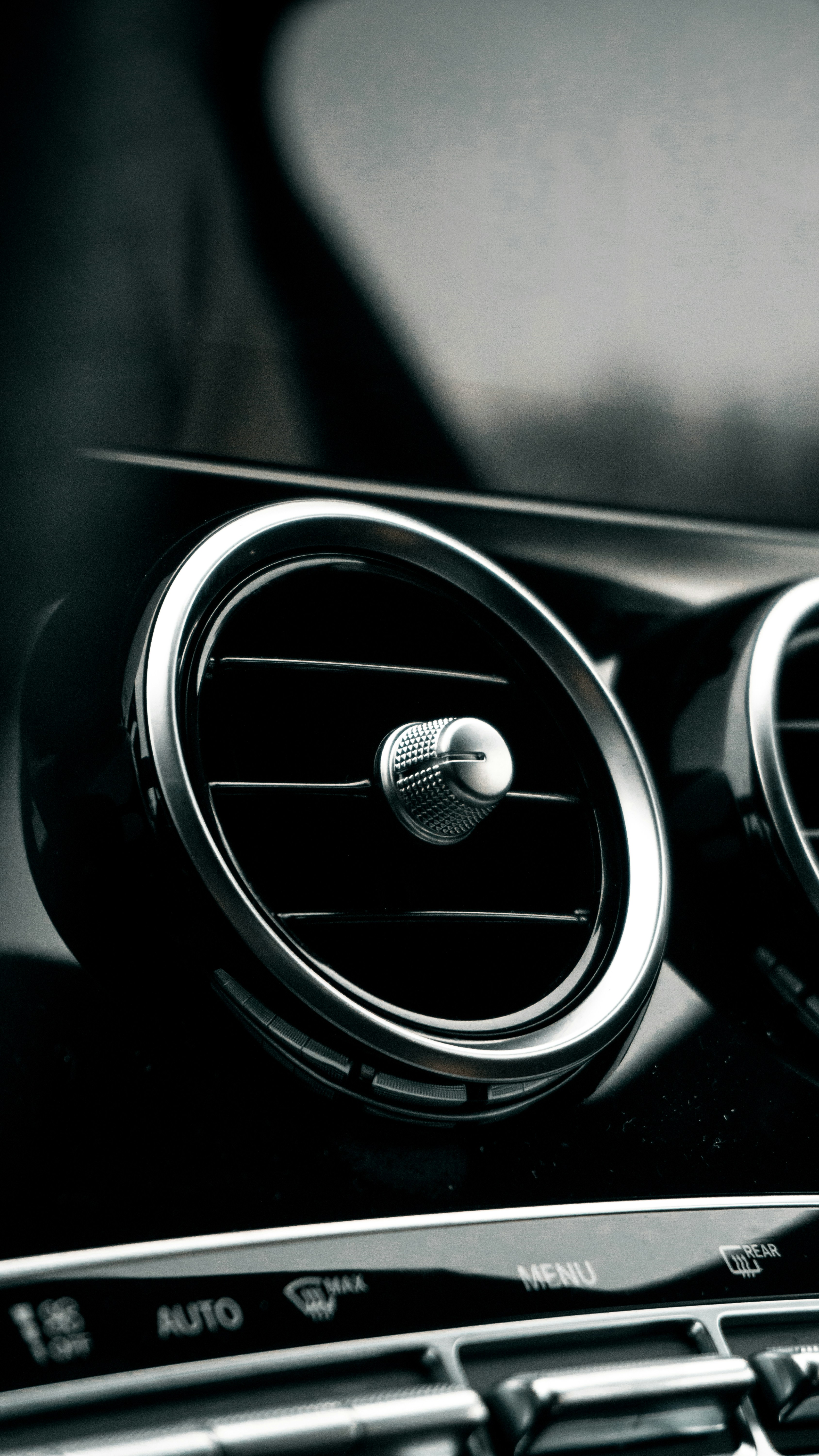 A car interior focusing on the air conditioning vents with a sleek, metallic appearance. The dashboard controls are visible at the bottom, featuring buttons and a 'MENU' label.