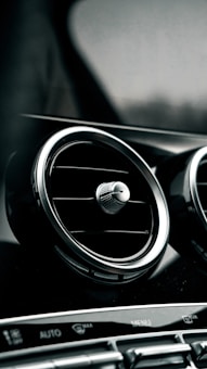 A car interior focusing on the air conditioning vents with a sleek, metallic appearance. The dashboard controls are visible at the bottom, featuring buttons and a 'MENU' label.