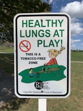 A sign displays the message 'HEALTHY LUNGS AT PLAY' in large green letters, with a no-smoking symbol and a smaller text indicating 'This is a tobacco-free zone.' It includes a cartoon illustration of children playing on a seesaw in a park setting.