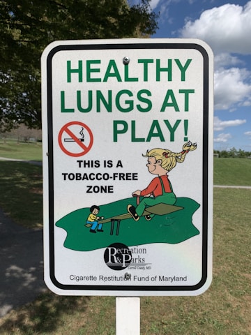 A sign displays the message 'HEALTHY LUNGS AT PLAY' in large green letters, with a no-smoking symbol and a smaller text indicating 'This is a tobacco-free zone.' It includes a cartoon illustration of children playing on a seesaw in a park setting.