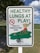 A sign displays the message 'HEALTHY LUNGS AT PLAY' in large green letters, with a no-smoking symbol and a smaller text indicating 'This is a tobacco-free zone.' It includes a cartoon illustration of children playing on a seesaw in a park setting.