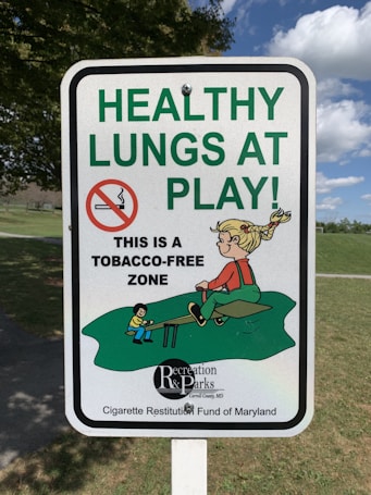 A sign displays the message 'HEALTHY LUNGS AT PLAY' in large green letters, with a no-smoking symbol and a smaller text indicating 'This is a tobacco-free zone.' It includes a cartoon illustration of children playing on a seesaw in a park setting.