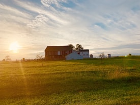 A serene rural landscape featuring a rustic barn and a few smaller farm buildings, set against a vibrant sunset sky with scattered clouds. The foreground displays an expanse of lush, green grass, while the golden sunlight casts a warm glow over the entire scene.