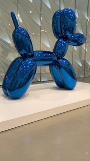 A large, shiny blue sculpture resembling a balloon dog is prominently displayed on a white platform. The reflective surface shows intricate highlights and shadows, and the background features a modern, geometric wall design.
