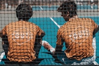 Two young people wearing orange shirts with 'YOUTH' printed on the back sit together on a sports court, viewed through a black netting. The court is blue, and one of them has a leg stretched out while they appear to be engaged in conversation or rest.