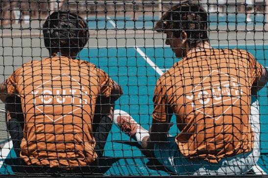 Two young people wearing orange shirts with 'YOUTH' printed on the back sit together on a sports court, viewed through a black netting. The court is blue, and one of them has a leg stretched out while they appear to be engaged in conversation or rest.