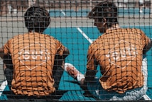 Two young people wearing orange shirts with 'YOUTH' printed on the back sit together on a sports court, viewed through a black netting. The court is blue, and one of them has a leg stretched out while they appear to be engaged in conversation or rest.
