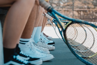 Several pairs of legs dressed in athletic shoes are seen from the knees down, positioned alongside a row of tennis rackets, indicating preparation for a tennis game on an outdoor court.