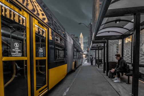 A city bus parked at a bus stop on a dimly lit street. The bus, primarily yellow and gray with a sign indicating fare payments are temporarily suspended, reflects the urban surroundings. On the right, a man sits under a covered bus stop shelter, waiting and staring at his phone. In the background, there are tall buildings, including a distinctively shaped skyscraper.
