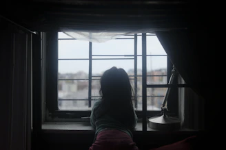 A quiet moment captured of a child looking out a window, with a distant view of a war-torn neighborhood.