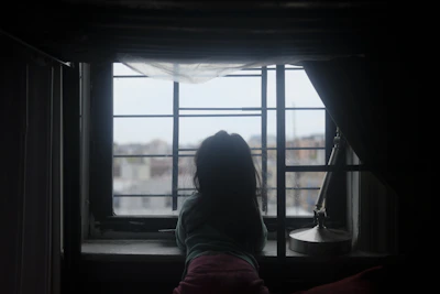 A quiet moment captured of a child looking out a window, with a distant view of a war-torn neighborhood.