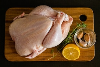 A whole raw chicken placed on a wooden cutting board alongside a halved lemon, a small glass jar containing garlic cloves and peppercorns, and sprigs of fresh herbs. The board is set against a dark background.