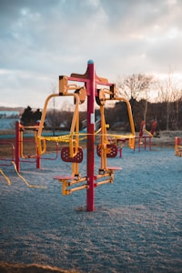 An outdoor gym equipment set in a park with caution tape wrapped around it. The equipment is primarily red and yellow, situated on a frosty ground with trees and cloudy skies in the background. The park appears to be closed or restricted.