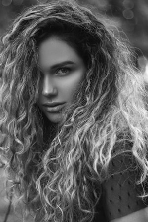 A black and white portrait of a person with long, curly hair cascading across their shoulders. The lighting accentuates their facial features, creating a dramatic contrast and adding depth to the curly texture. The background is softly blurred, drawing attention to the expression and curly hair.