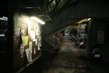 A dimly lit outdoor bulletin board with various papers and notices attached to it. The scene is set in an alley with a narrow walkway that leads to a stairway on the left. Several parked scooters are visible on the right side, and overhead are some signs with text in a non-Latin script. Trees can be seen in the background, adding to the slightly dark and shaded atmosphere.