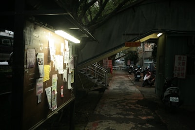 A dimly lit outdoor bulletin board with various papers and notices attached to it. The scene is set in an alley with a narrow walkway that leads to a stairway on the left. Several parked scooters are visible on the right side, and overhead are some signs with text in a non-Latin script. Trees can be seen in the background, adding to the slightly dark and shaded atmosphere.