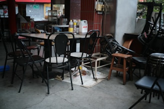 A dining area with a clutter of chairs stacked around a table. The table is covered with bottles and a small carton, surrounded by various unmatched chairs. There are cleaning supplies and signs of an eatery setting, with red tiles and a glimpse of a kitchen in the background.