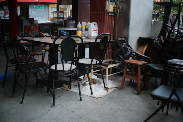 A dining area with a clutter of chairs stacked around a table. The table is covered with bottles and a small carton, surrounded by various unmatched chairs. There are cleaning supplies and signs of an eatery setting, with red tiles and a glimpse of a kitchen in the background.